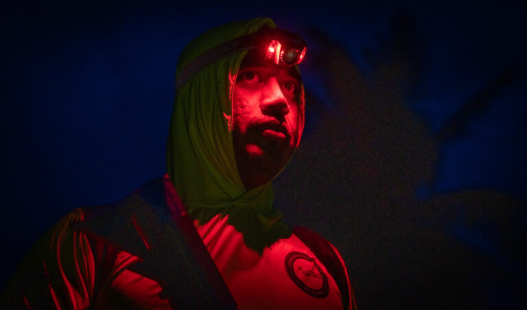 John Reynold wearing a lime green hood and a red headlamp looking upward at night.