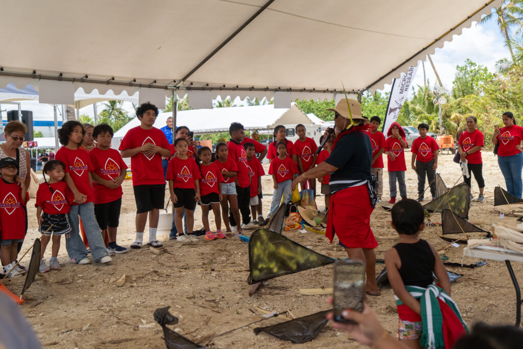 A group of children gather under a large white event tent on a sandy ground. . On the right, Master Navigator Larry Raigatel speaks to the group.