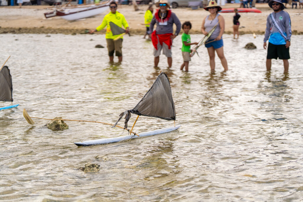 A close-up shot focuses on a small, hand-crafted galaide’ sailing in shallow ocean water.