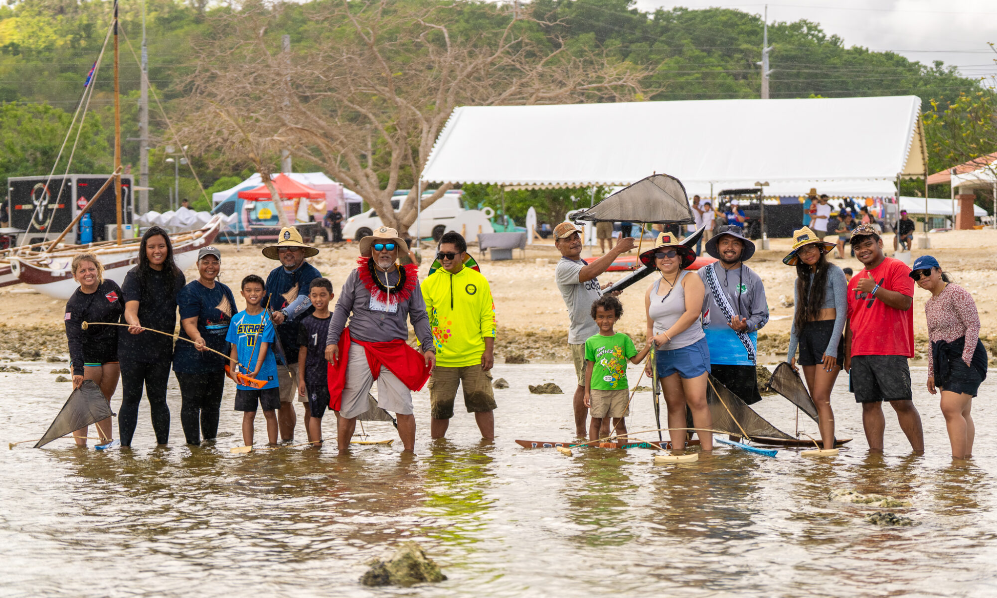 Students and their families standing knee-deep in calm, shallow ocean water, standing near small model versions of galaide’ or flying proas.