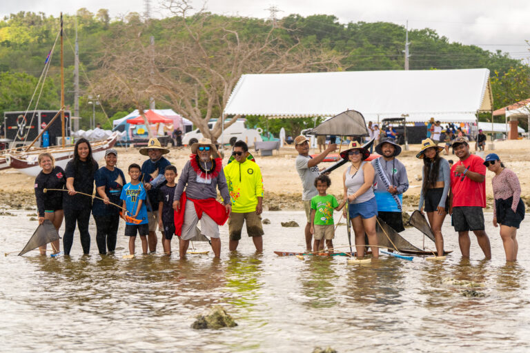 Students and their families standing knee-deep in calm, shallow ocean water, standing near small model versions of galaide’ or flying proas.