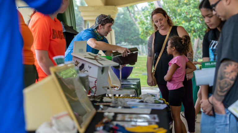 An outdoor educational outreach table where a presenter holds out a container to show a young girl and a woman, while other attendees gather around to learn about brown treesnakes.