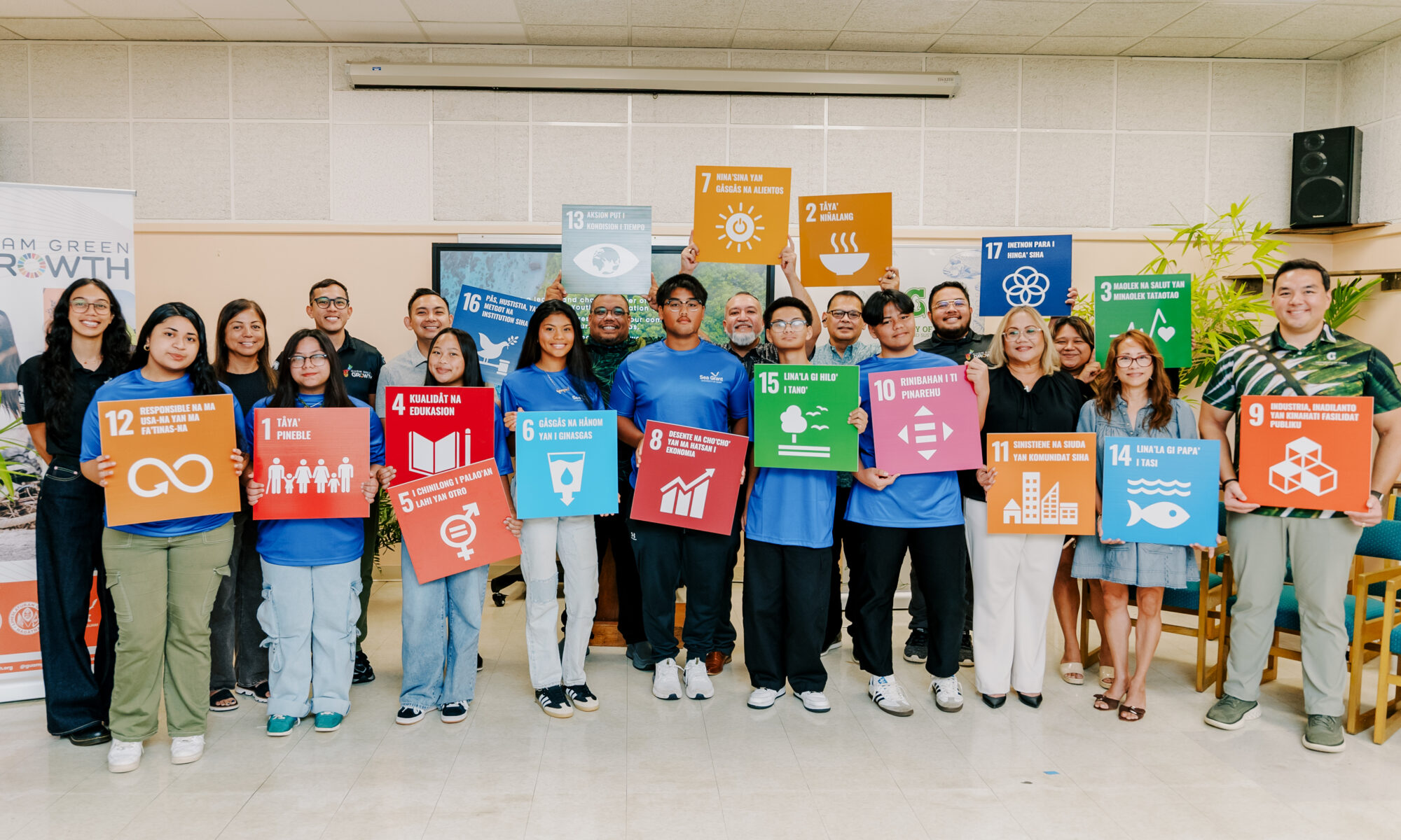 A large group of youth conservation corps members and leadership pose indoors, holding up colorful square signs with various icons and Chamoru text representing local sustainability and conservation goals.