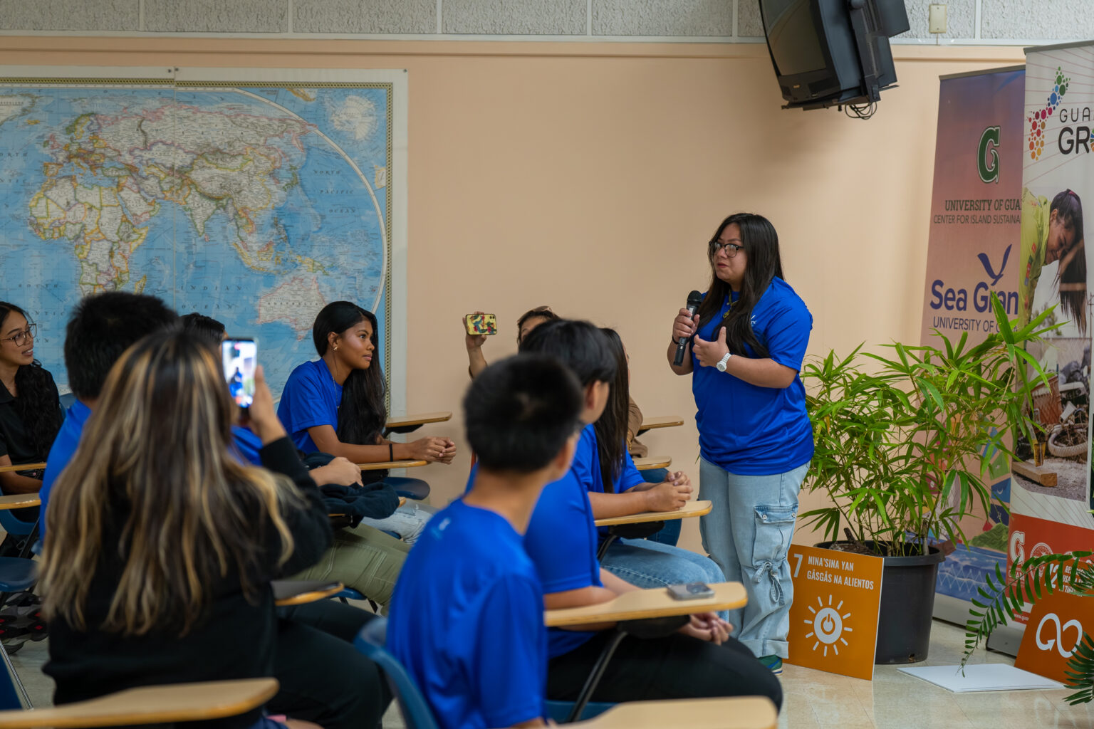 A young female student wearing a blue t-shirt stands and speaks into a microphone during a presentation inside a classroom setting at the UOG RFK Library, while other seated youth conservation corps members listen attentively and record with smartphones.
