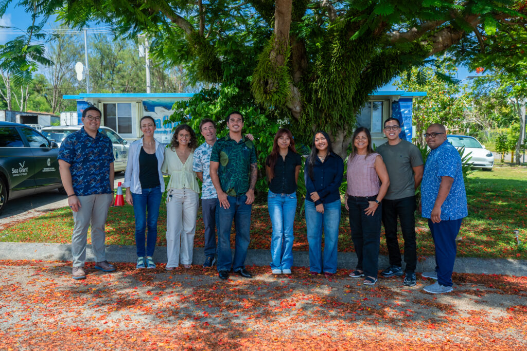 A group of ten people, including student fellows and UOG Sea Grant staff, pose for a photo under a large tree surrounded by fallen red blossoms outside a Sea Grant building.