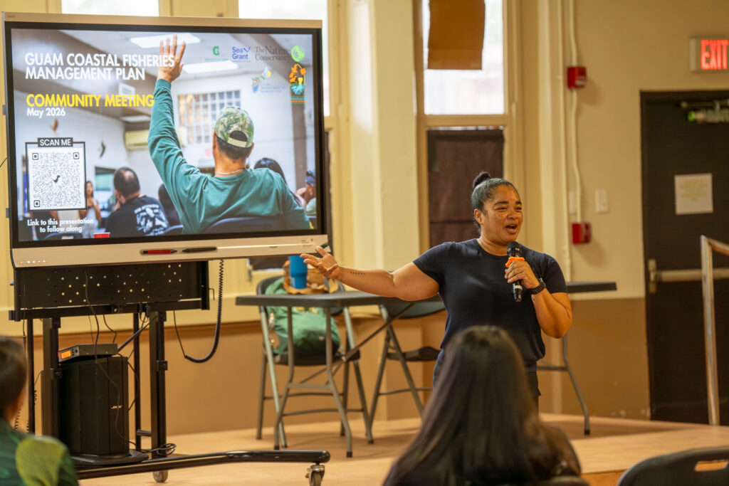 Guam Department of Agriculture Director Chelsa Muna stands indoors speaking into a microphone, gesturing toward a large digital display screen that reads "Guam Coastal Fisheries Management Plan Community Meeting" next to an audience member.