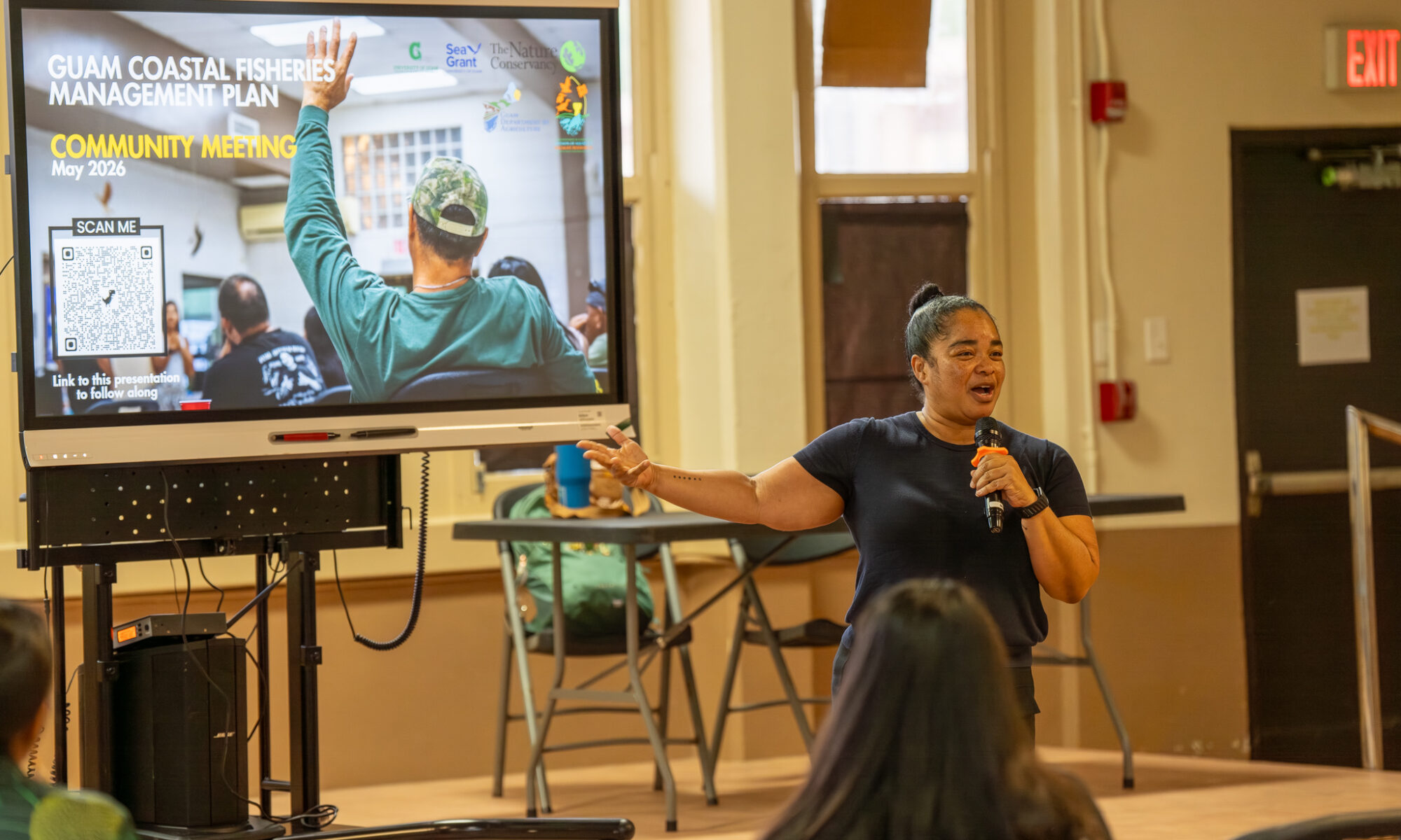 Guam Department of Agriculture Director Chelsa Muna stands indoors speaking into a microphone, gesturing toward a large digital display screen that reads "Guam Coastal Fisheries Management Plan Community Meeting" next to an audience member.