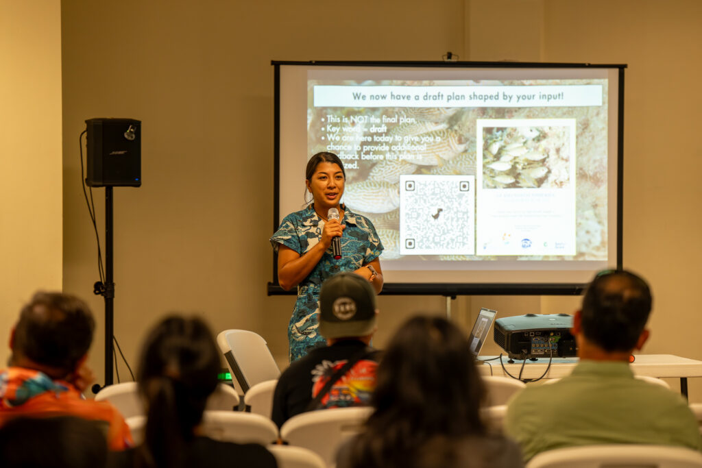 Leilani Sablan Naden stands at the front of a room speaking into a microphone during a community meeting, with a projector screen behind her displaying information about a draft plan to a seated audience.