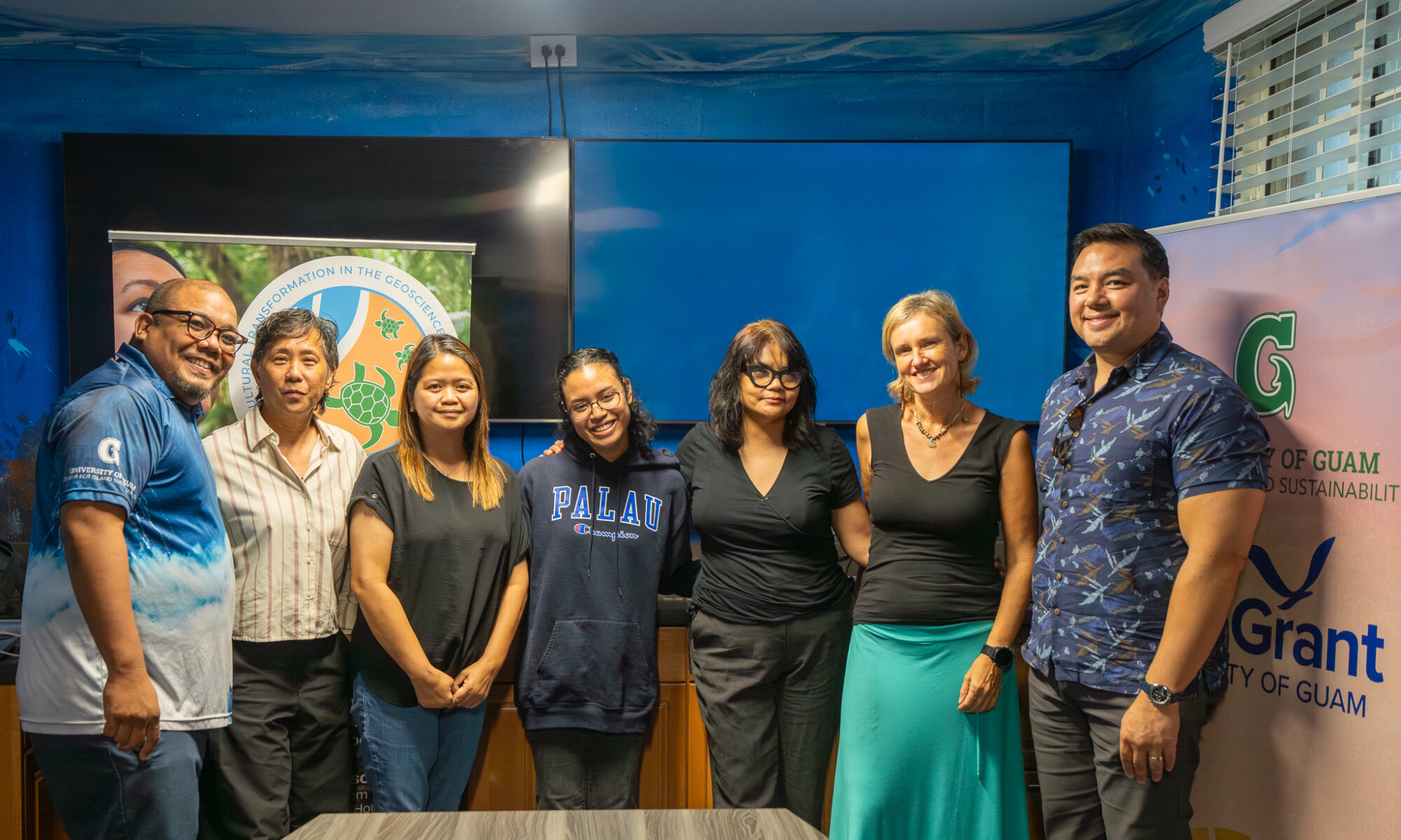 A group of seven people, including Navigating Home Fellow Kira Reklai, stand smiling side-by-side indoors for a photo following a final fellowship presentation.