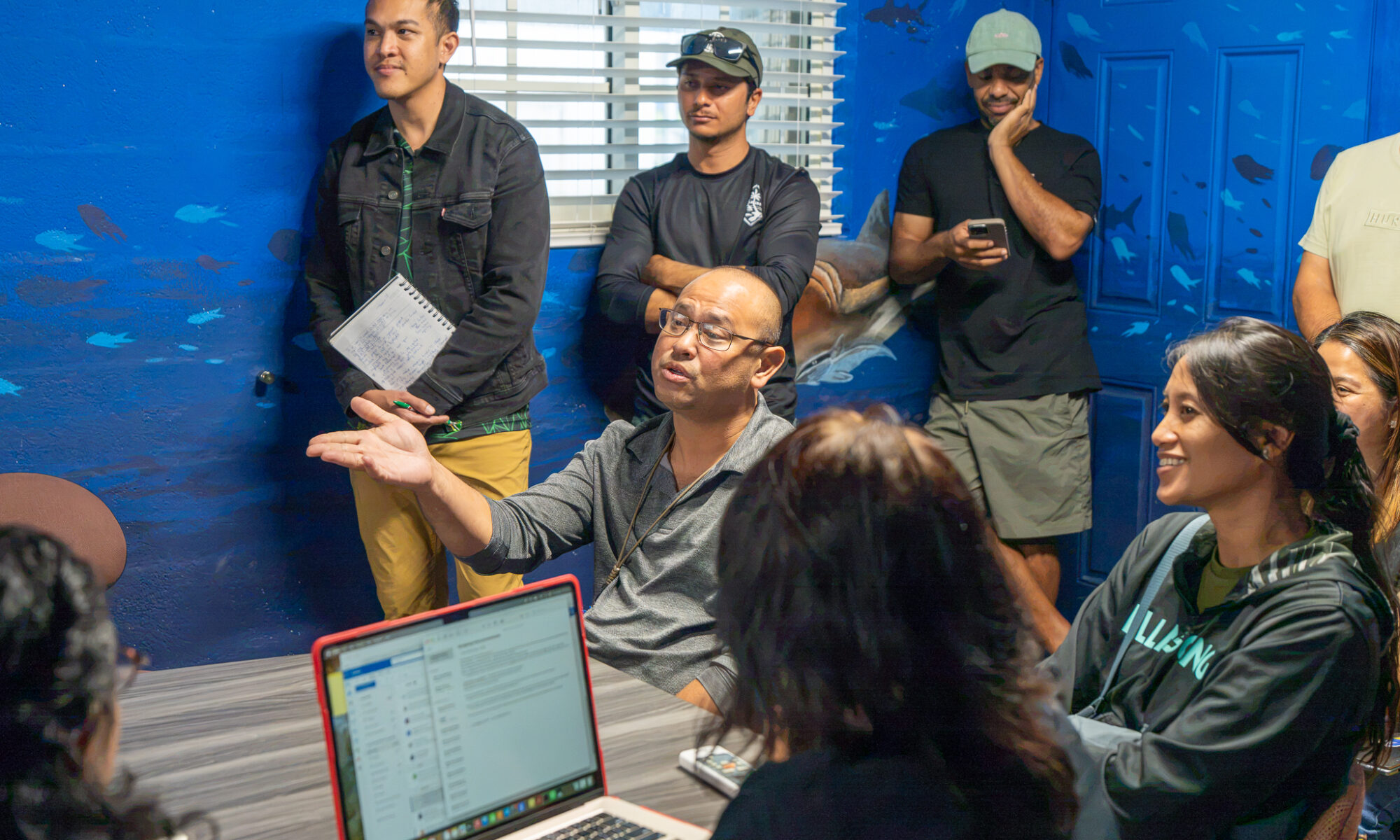 Michael Duenas, Guam Department of Agriculture Fisheries Supervisor, sits at a table and gestures with his hand while speaking to a group of attendees gathered in a room with blue, ocean-themed walls.