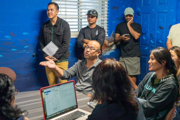 Michael Duenas, Guam Department of Agriculture Fisheries Supervisor, sits at a table and gestures with his hand while speaking to a group of attendees gathered in a room with blue, ocean-themed walls.