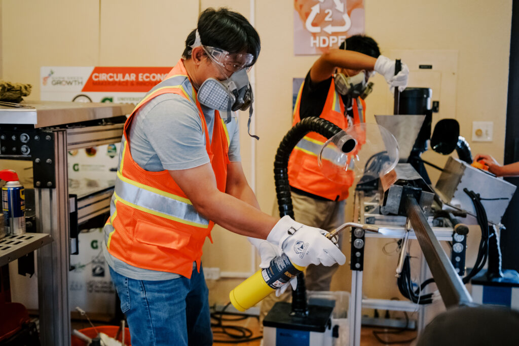 Dr. Ernesto J. Guades, wearing protective gear, uses a blowtorch on recyclable plastics at the G3 Makerspace while a student operates machinery in the background.