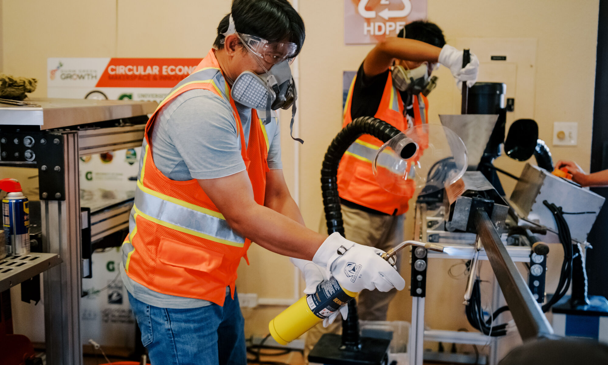 Dr. Ernesto J. Guades, wearing protective gear, uses a blowtorch on recyclable plastics at the G3 Makerspace while a student operates machinery in the background.