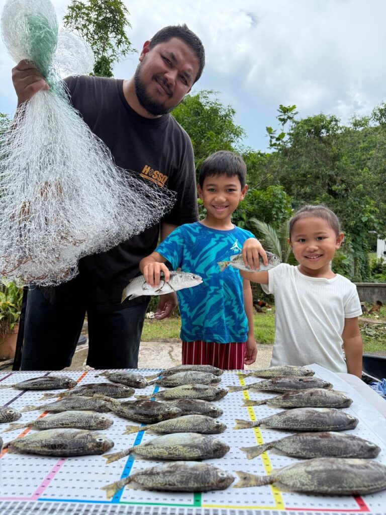 A fisher and his kids pose in front of a Guihan for Guåhan catch mat lined with reef fish.