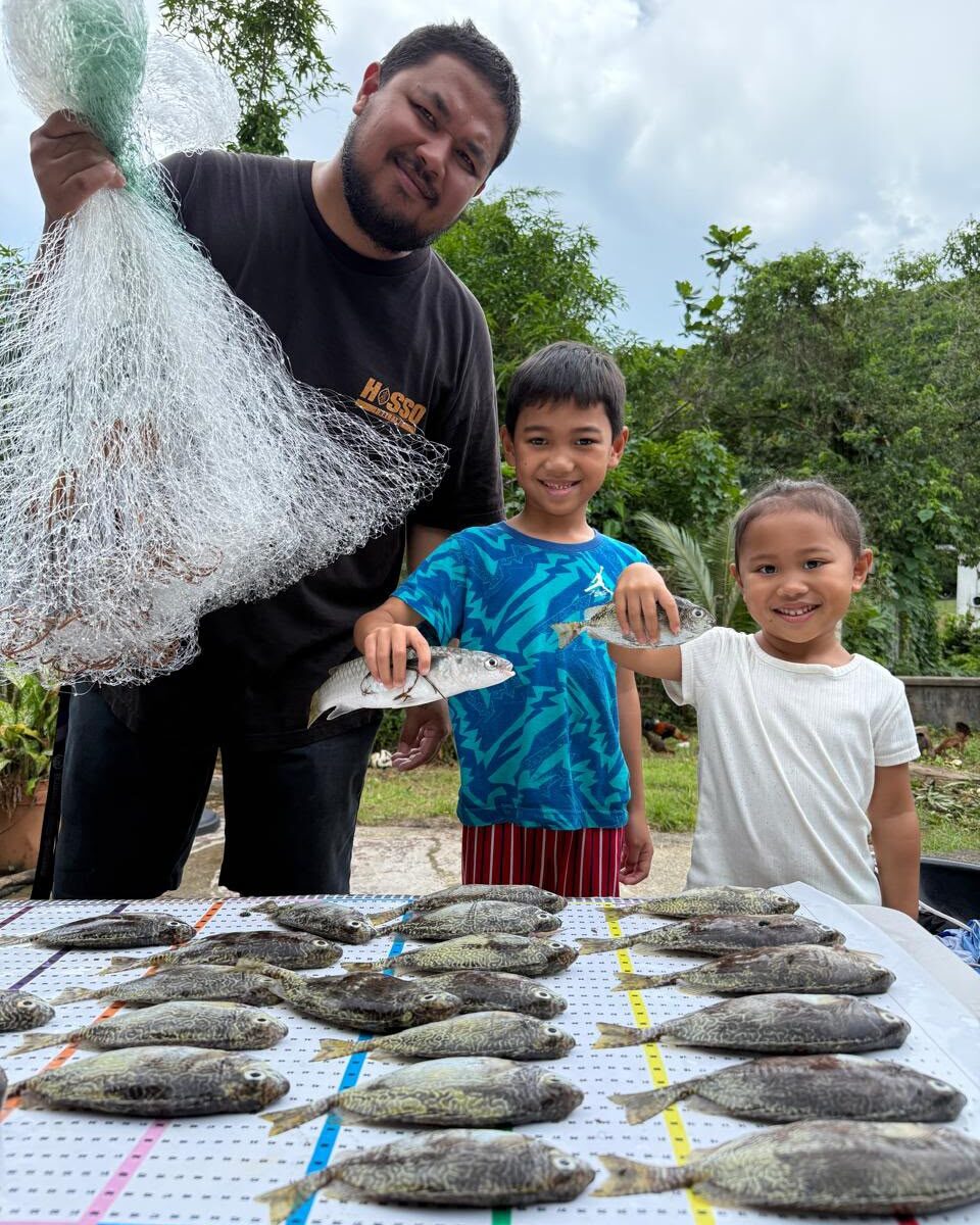 A fisher and his kids pose in front of a Guihan for Guåhan catch mat lined with reef fish.